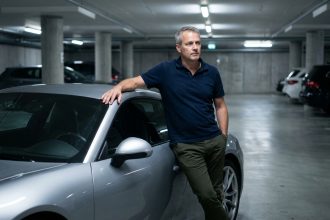 Homme passionné de Porsche appuyé contre sa 911 argentée dans un garage béton, réfléchissant à son assurance auto